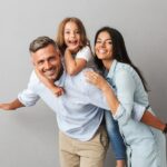 A young child sits on their dad's back while a mom leans in and places her hand on his shoulder. Everyone is smiling.