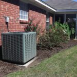 A large, gray air conditioning unit sits on a concrete slab in the backyard of a brick yard. The unit is next to a rose bush.