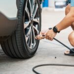 An individual in a blue T-shirt, white shorts, and black sandals is using an air pump to inflate a car tire.