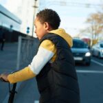 A child in a puffer vest crossing a busy street while riding a black scooter. Cars wait for him to pass.