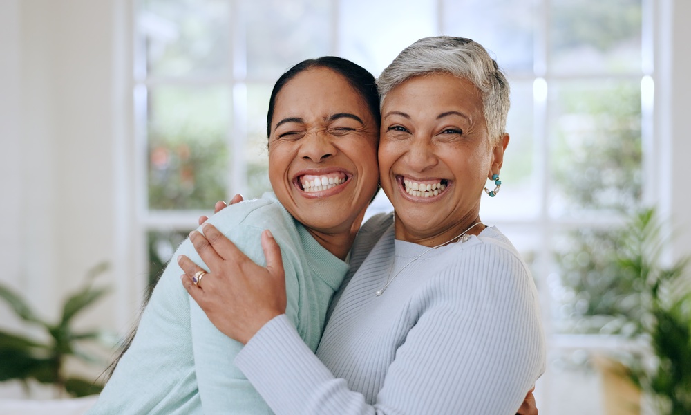 An adult daughter and her aging mother embracing and grinning broadly in front of a living room window.