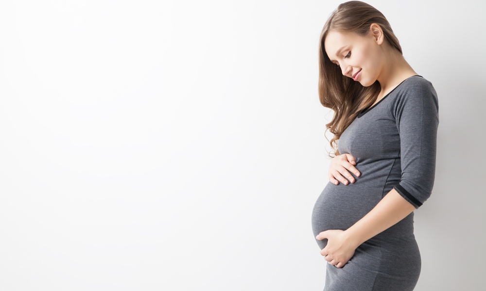 A pregnant woman in a gray dress stands against a white background. She is caressing her belly and staring at it lovingly.