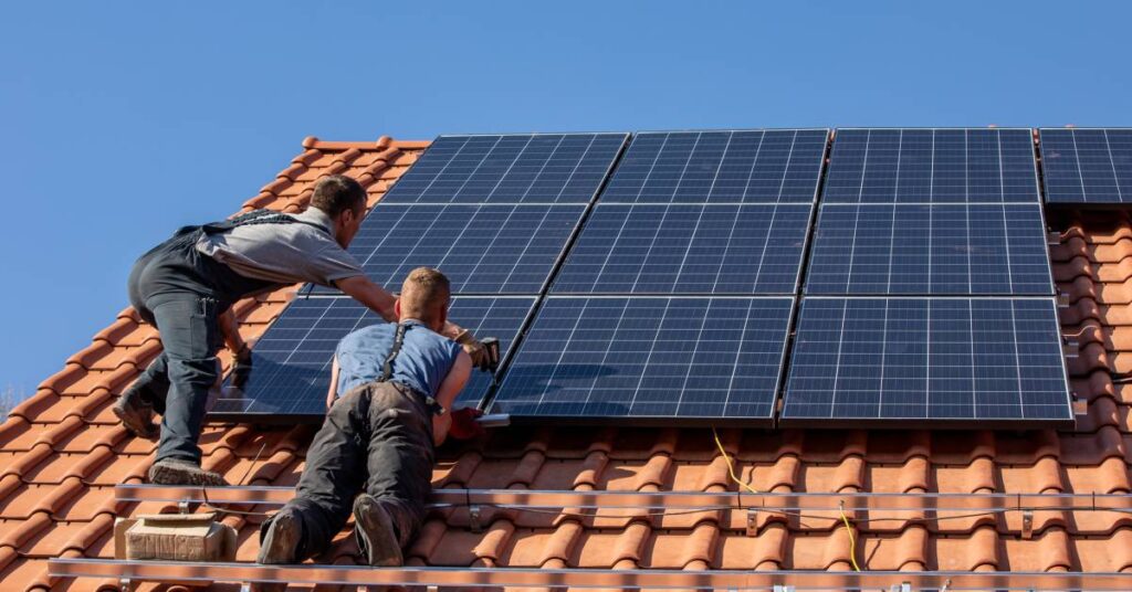 Two men wearing overalls and T-shirts install solar panels on a stucco roof with a blue sky in the background.