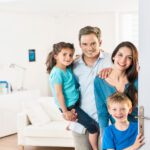 A family stands behind an open front door, smiling toward the camera. The background is living room furniture and decor.