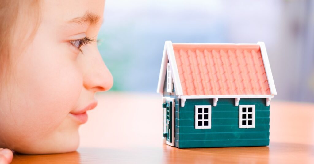 A child rests their chin on the surface of a wooden table and looks straight ahead at a model of a house.