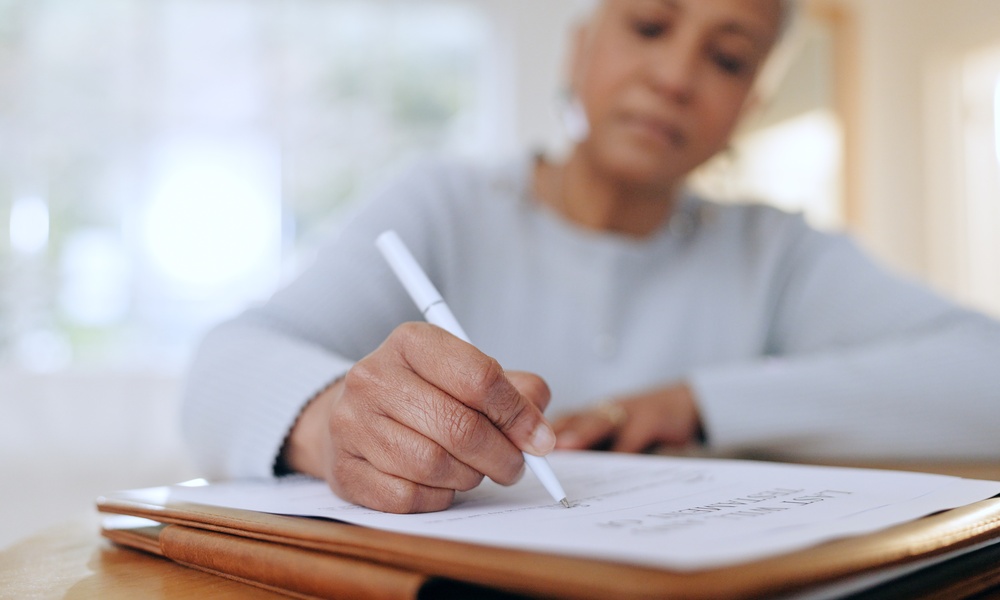 An older woman is looking down at a document on top of a leather folder, signing the document with a white pen.