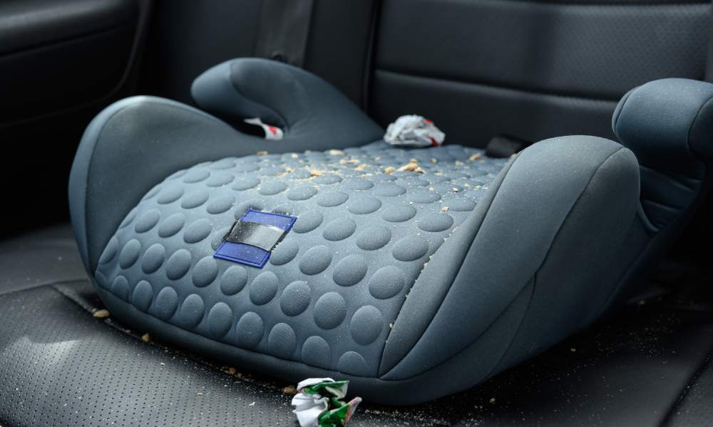 A child's dark gray booter seat in the backseat of a car. Crumbs and wrappers from snacks cover the booster seat.