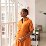 A pregnant woman wearing a bright orange dress smiles and cradles her belly while looking out the window.