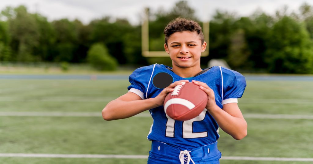 A young boy stands on a football field wearing a blue football jersey and holding a football. He is smiling.