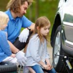A dad sitting with his kids looking at a tire on their car. The car is jacked up, and one of the kids is sitting on a spare tire.