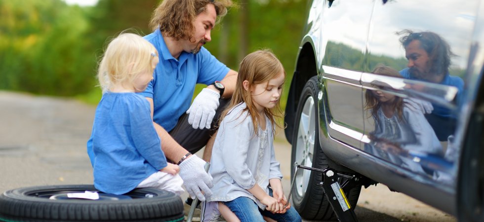 A dad sitting with his kids looking at a tire on their car. The car is jacked up, and one of the kids is sitting on a spare tire.