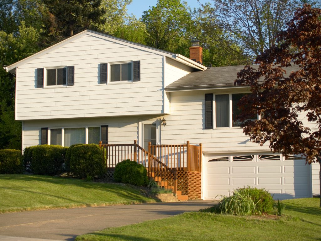 A white split-level home contains windows with black shutters, a white garage door, and a wooden front deck.