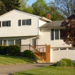 A white split-level home contains windows with black shutters, a white garage door, and a wooden front deck.