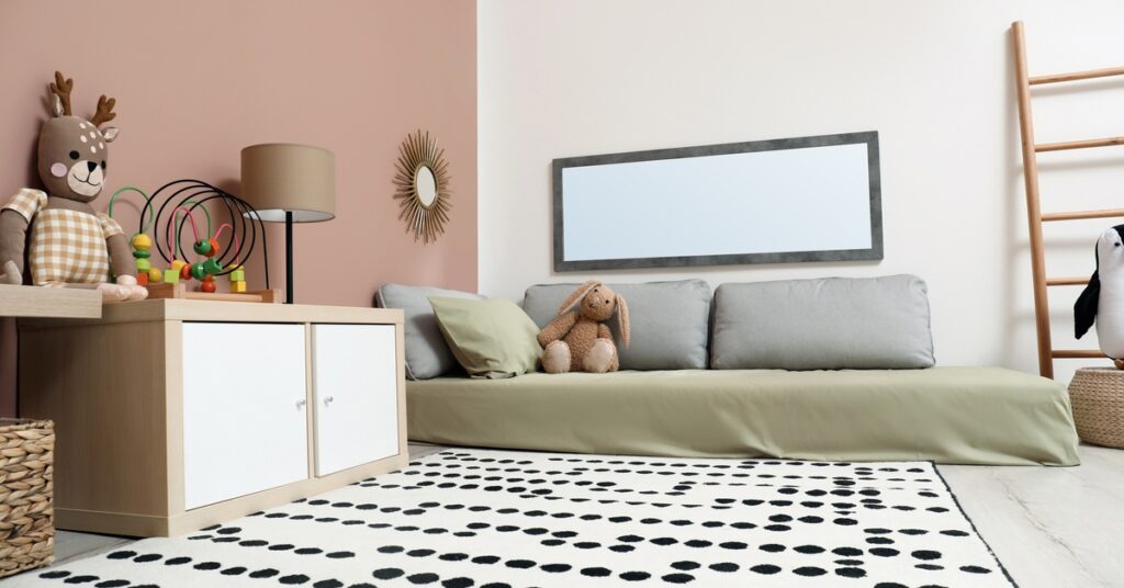 Gray marble floors are covered with a dotted rug near a bed with olive bedding, stuffed animals, and wicker storage.