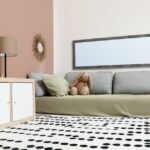 Gray marble floors are covered with a dotted rug near a bed with olive bedding, stuffed animals, and wicker storage.