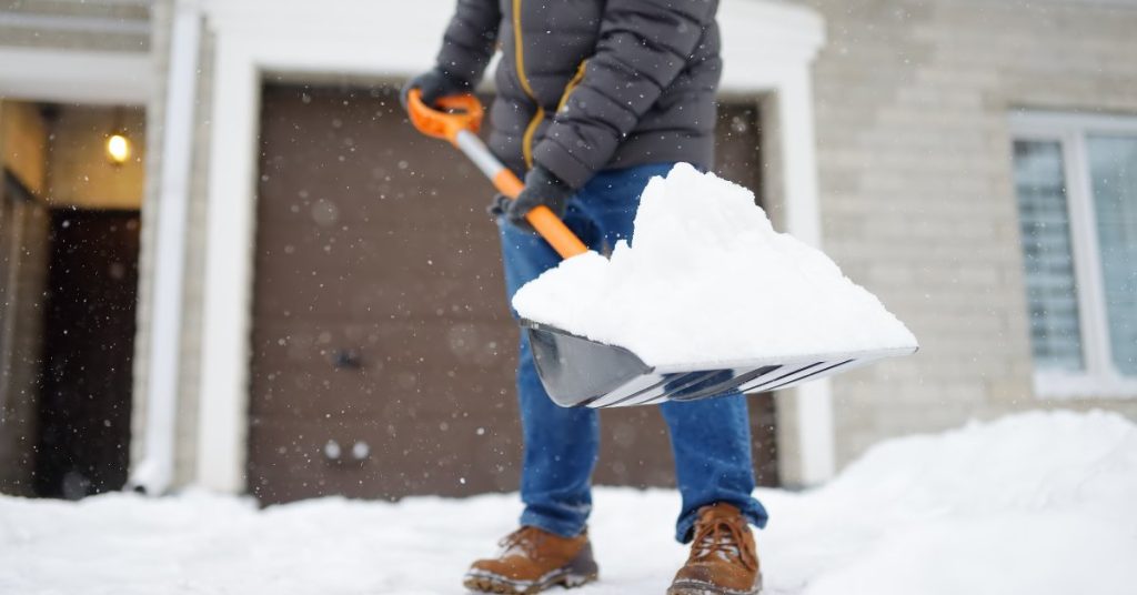 A person in a puffer coat, jeans, and boots is shoveling snow on the driveway in front of a beige house.