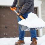 A person in a puffer coat, jeans, and boots is shoveling snow on the driveway in front of a beige house.