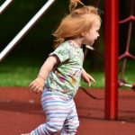A red-headed toddler running through a playground with red rubber surfacing. A climbing net is behind her.