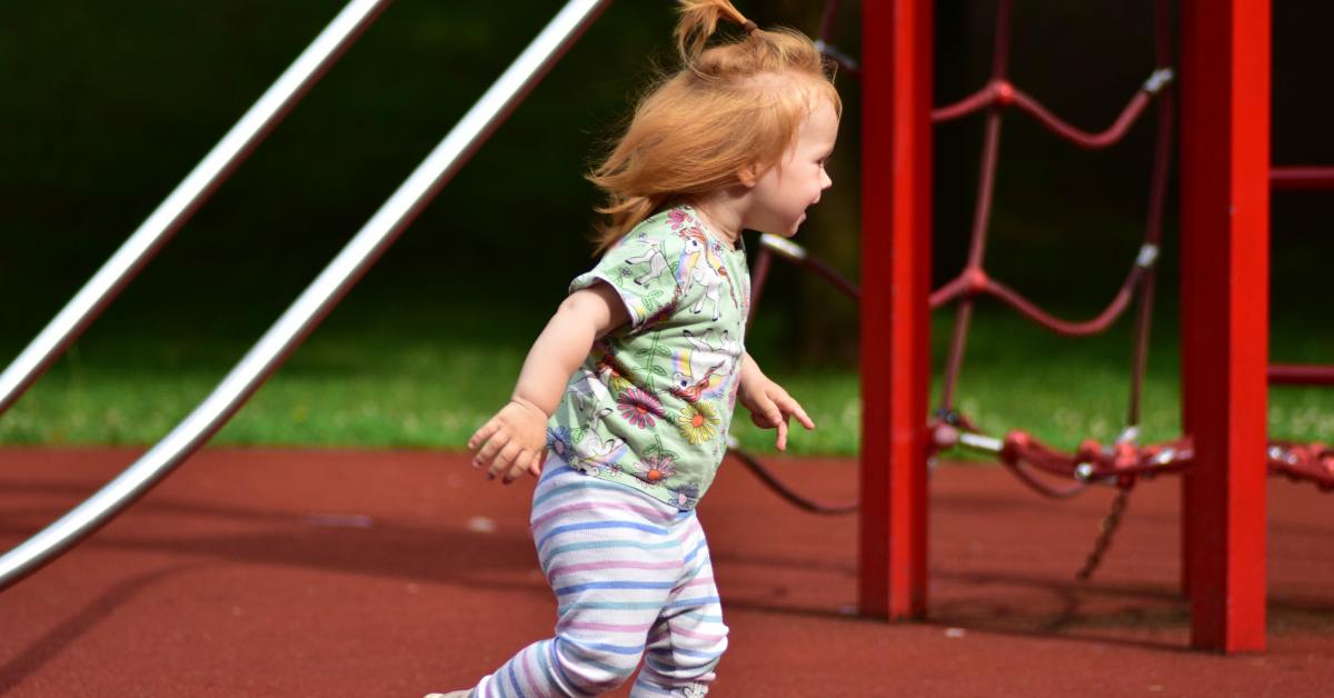 A red-headed toddler running through a playground with red rubber surfacing. A climbing net is behind her.