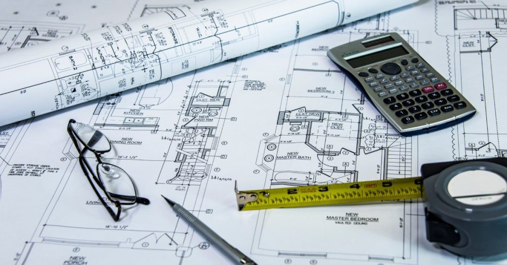 Blueprints are spread out on a table while a measuring tape, a calculator, a pen, and a pair of glasses sit on top.