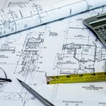 Blueprints are spread out on a table while a measuring tape, a calculator, a pen, and a pair of glasses sit on top.