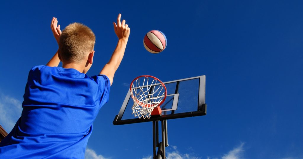 A boy wearing a blue shirt with blonde hair is shooting a blue, red, and white basketball toward a basketball hoop.