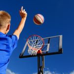 A boy wearing a blue shirt with blonde hair is shooting a blue, red, and white basketball toward a basketball hoop.