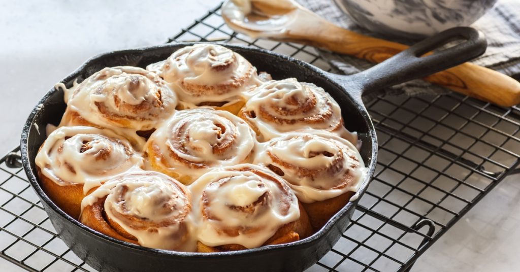 A 12 inch cast iron pan sits on a cooling rack after baking cinnamon rolls. The icing is melting into the folds.