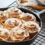 A 12 inch cast iron pan sits on a cooling rack after baking cinnamon rolls. The icing is melting into the folds.
