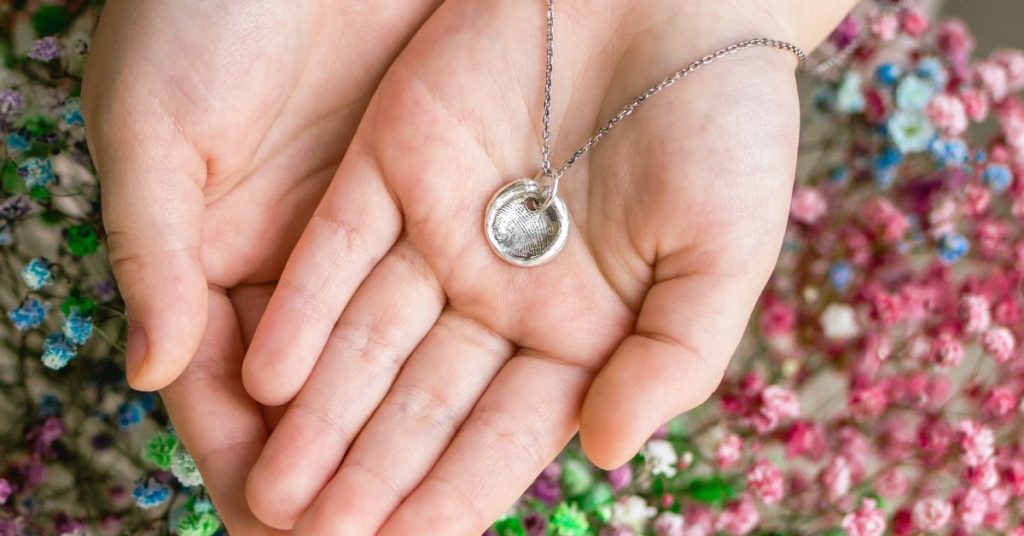 A small silver necklace pendant with the impression of someone's fingerprint sitting in a person's cupped palms.
