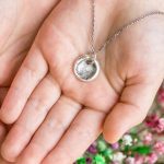 A small silver necklace pendant with the impression of someone's fingerprint sitting in a person's cupped palms.