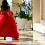 A young woman in a voluminous red gown with ruffles is seen from behind, walking in an elegant outdoor corridor.