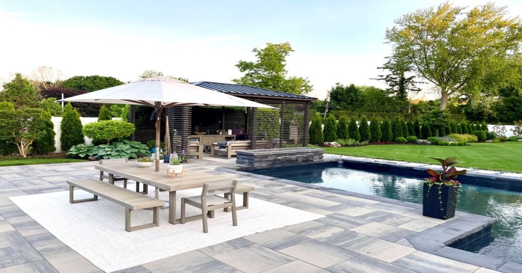 A high-end pool sits in a residential backyard surrounded by an outdoor gray tile. A pergola and a dining table are next to it.