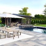A high-end pool sits in a residential backyard surrounded by an outdoor gray tile. A pergola and a dining table are next to it.