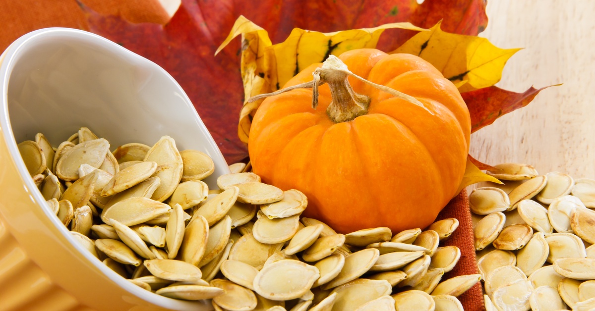 A ceramic bowl filled with toasted pumpkin seeds, with some spilling out, surrounded by a mini pumpkin and autumn leaves.