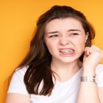 A brown-haired girl in a white t-shirt with braces frowning and clenching her fist near her face against a yellow background.