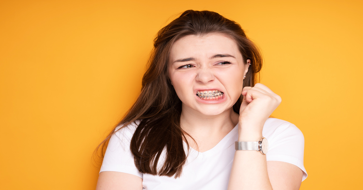 A brown-haired girl in a white t-shirt with braces frowning and clenching her fist near her face against a yellow background.