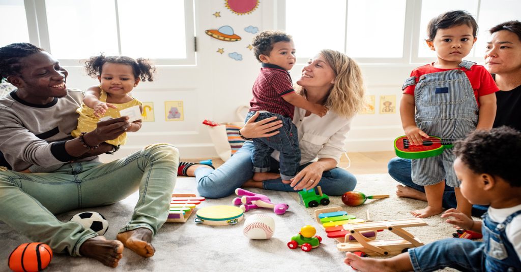 A diverse group of adults and children play on a carpet with toys in a bright, colorful child-friendly room