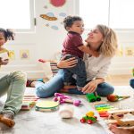 A diverse group of adults and children play on a carpet with toys in a bright, colorful child-friendly room