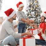A family of four wears Santa hats as they sit on the floor, passing gifts around. They're all wearing jeans and gray tops.