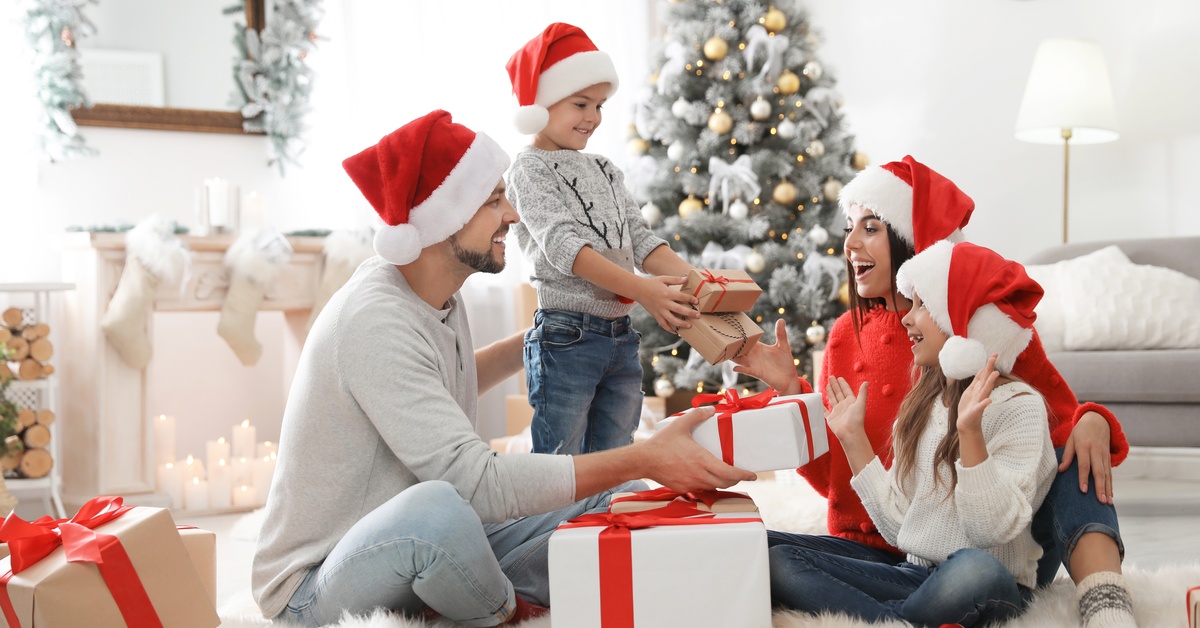 A family of four wears Santa hats as they sit on the floor, passing gifts around. They're all wearing jeans and gray tops.