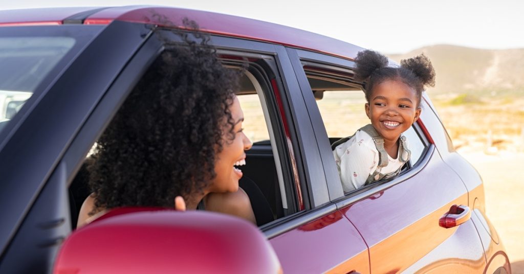 A Black woman in the driver's seat of a red car looks out of her window at a smiling child while parked.