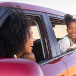 A Black woman in the driver's seat of a red car looks out of her window at a smiling child while parked.