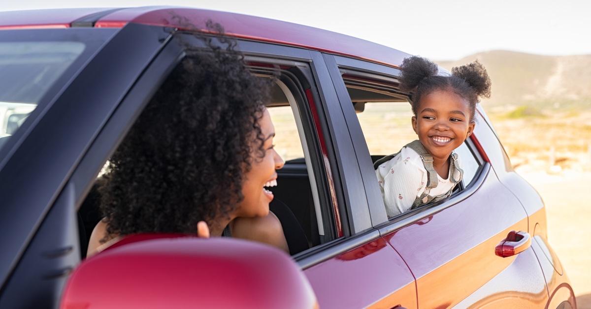 A Black woman in the driver's seat of a red car looks out of her window at a smiling child while parked.