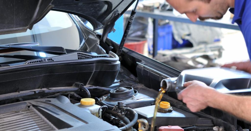 A brown-haired man in a blue shirt pours amber-colored liquid from a silver container into a vehicle engine.