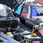 A brown-haired man in a blue shirt pours amber-colored liquid from a silver container into a vehicle engine.