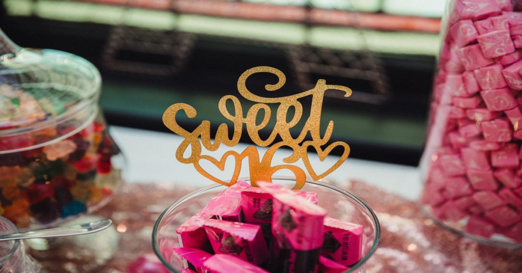 Three glass containers filled with candies, including gummy bears and Starbursts, with a "Sweet 16" topper in the center jar.