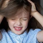 A close-up of a young girl with her hands in her long, brown hair, itching her scalp with an upset look on her face.