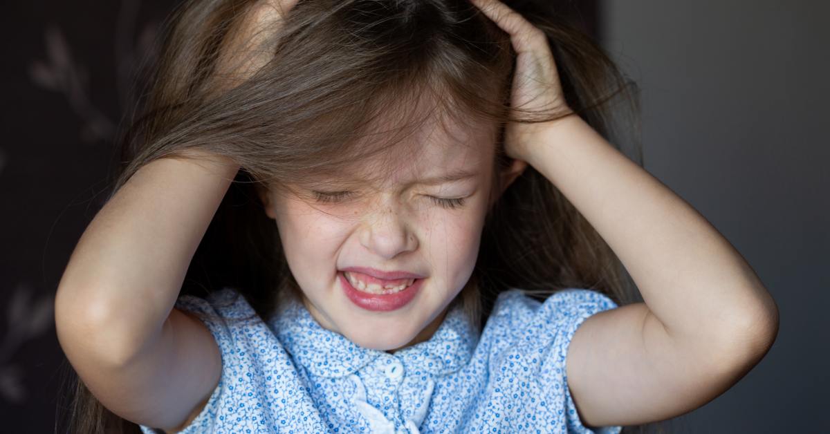 Home 1 A close-up of a young girl with her hands in her long, brown hair, itching her scalp with an upset look on her face.