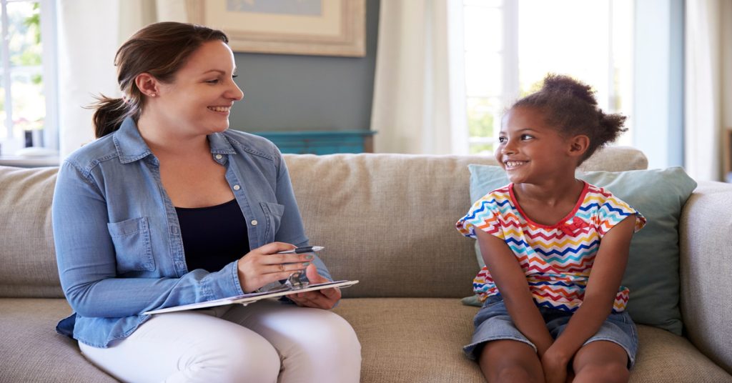 A therapist holds a clipboard during a session with a young child. They're both sitting on a couch and smiling at one another.